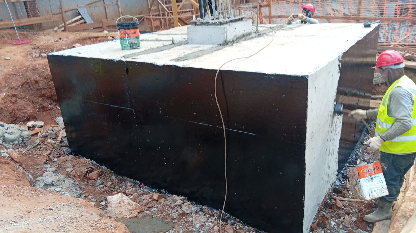 Equipe aplicando impermeabilização asfáltica em bloco de fundação na obra
